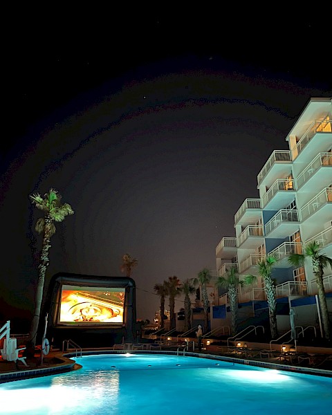 A nighttime outdoor movie setup by a swimming pool with an inflatable screen, surrounded by palm trees and a multi-story building with balconies.