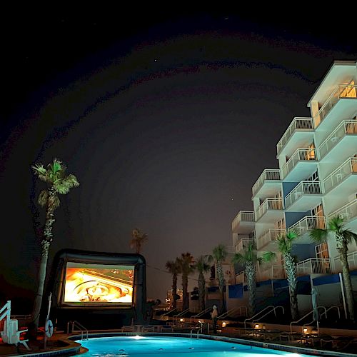 A nighttime outdoor movie setup by a swimming pool with an inflatable screen, surrounded by palm trees and a multi-story building with balconies.