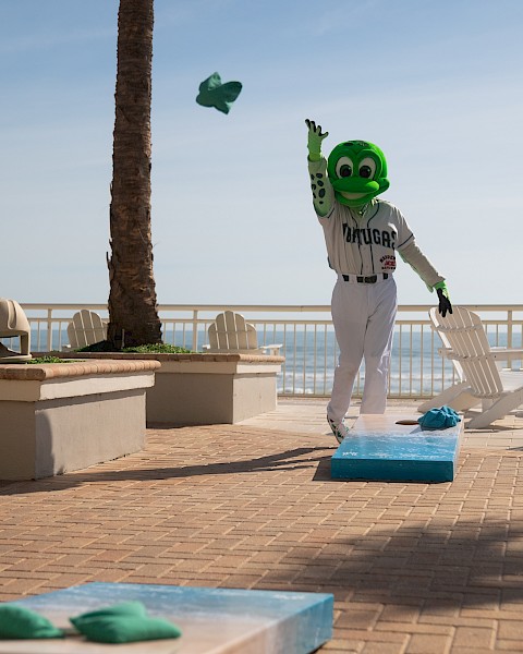 A person in a green mascot costume is playing a game of cornhole outdoors near palm trees and a beach.