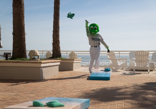 A person in a green mascot costume is playing a game of cornhole outdoors near palm trees and a beach.