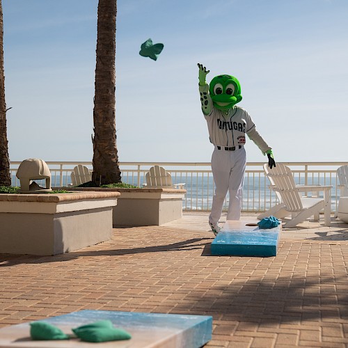 A person in a green mascot costume is playing a game of cornhole outdoors near palm trees and a beach.