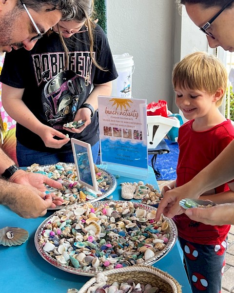 People are at a beach activity table collecting colorful seashells and rocks for keepsakes, smiling and enjoying the outdoor event.