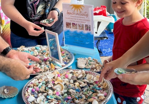 People are at a beach activity table collecting colorful seashells and rocks for keepsakes, smiling and enjoying the outdoor event.
