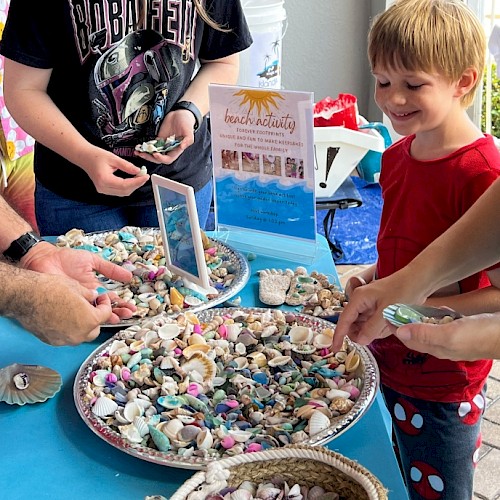 People are at a beach activity table collecting colorful seashells and rocks for keepsakes, smiling and enjoying the outdoor event.