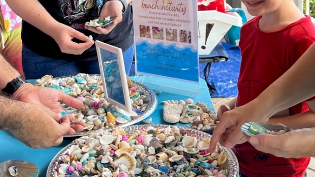People are at a beach activity table collecting colorful seashells and rocks for keepsakes, smiling and enjoying the outdoor event.