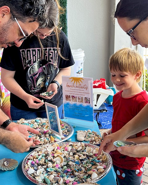 A group of people, including a child, looking at an assortment of colorful seashells and small trinkets displayed on plates set on a blue table.