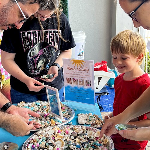 A group of people, including a child, looking at an assortment of colorful seashells and small trinkets displayed on plates set on a blue table.