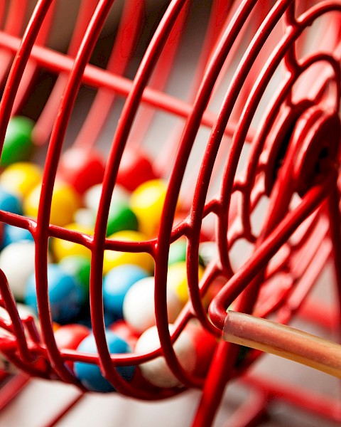 A close-up of a red metal bingo cage filled with colorful bingo balls, showing a handle for rotating the cage.