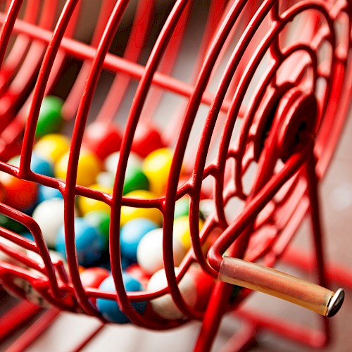 A close-up of a red metal bingo cage filled with colorful bingo balls, showing a handle for rotating the cage.