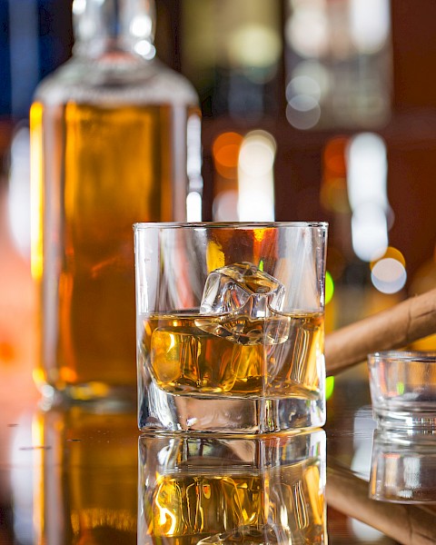 A glass of whiskey with ice, a cigar in an ashtray, and a bottle, set against a blurred background of colorful lights.