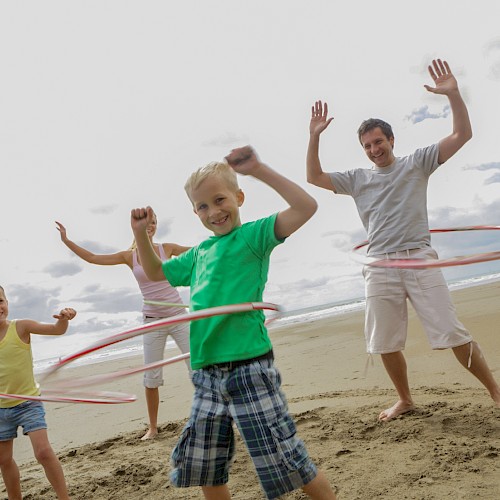 A family is having fun on the beach, hula hooping and smiling, with the ocean and cloudy sky in the background.