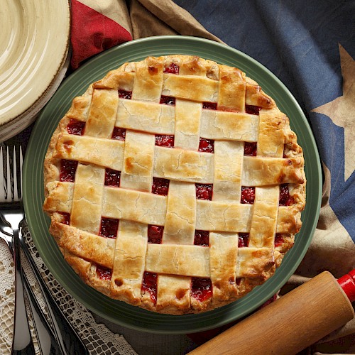 A cherry pie with a lattice crust on a green plate, surrounded by a Texas flag, utensils, a rolling pin, and a bowl of cherries ends the sentence.