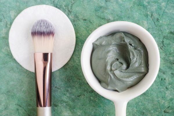 The image shows a makeup brush on a round pad next to a small white bowl of greyish-green facial clay mask on a green textured background.