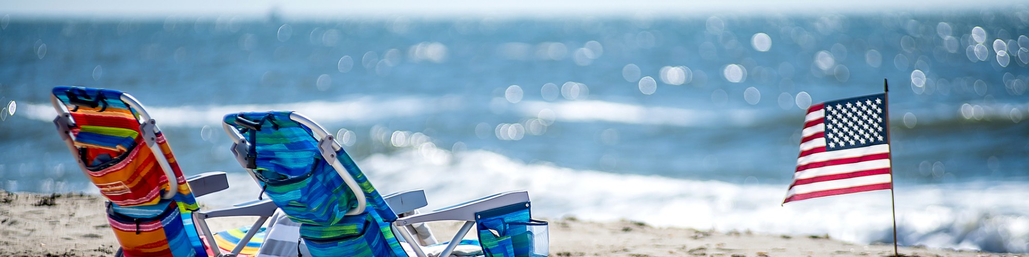 A sandy beach scene with two colorful beach chairs, a small table, and an American flag planted in the sand near the ocean waves.