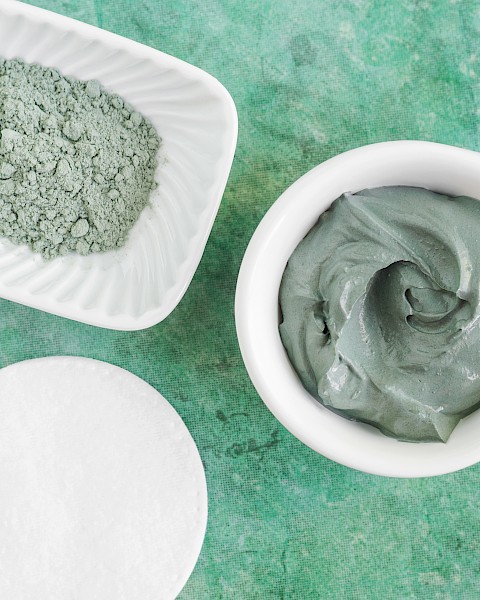 The image shows a bowl with gray clay paste, a dish with grayish powder, and a white cotton pad on a green textured background.
