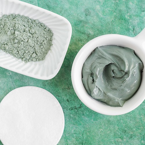 The image shows a bowl with gray clay paste, a dish with grayish powder, and a white cotton pad on a green textured background.