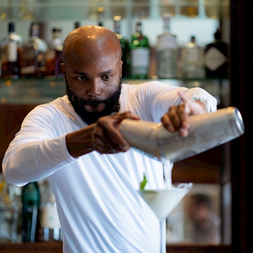 A bartender in a white shirt is pouring a drink from a shaker into a cocktail glass, with a bar filled with various bottles in the background.