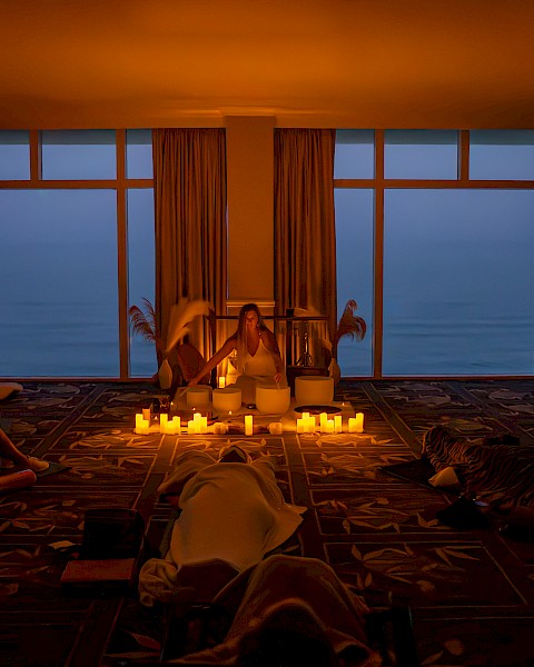 A dimly lit room with candles, people relaxing on the floor, and large windows showing an evening or early morning view outside.