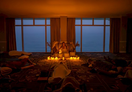 A dimly lit room with candles, people relaxing on the floor, and large windows showing an evening or early morning view outside.