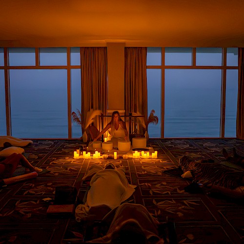 A dimly lit room with candles, people relaxing on the floor, and large windows showing an evening or early morning view outside.