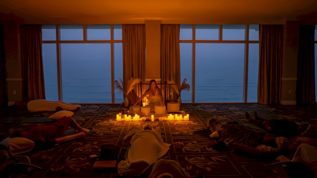 A dimly lit room with candles, people relaxing on the floor, and large windows showing an evening or early morning view outside.