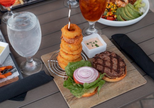 A meal with a burger, stacked onion rings, dipping sauce, a plate of salad, water, and iced tea on a table.