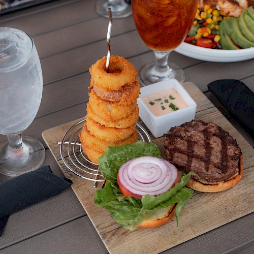 A meal with a burger, stacked onion rings, dipping sauce, a plate of salad, water, and iced tea on a table.