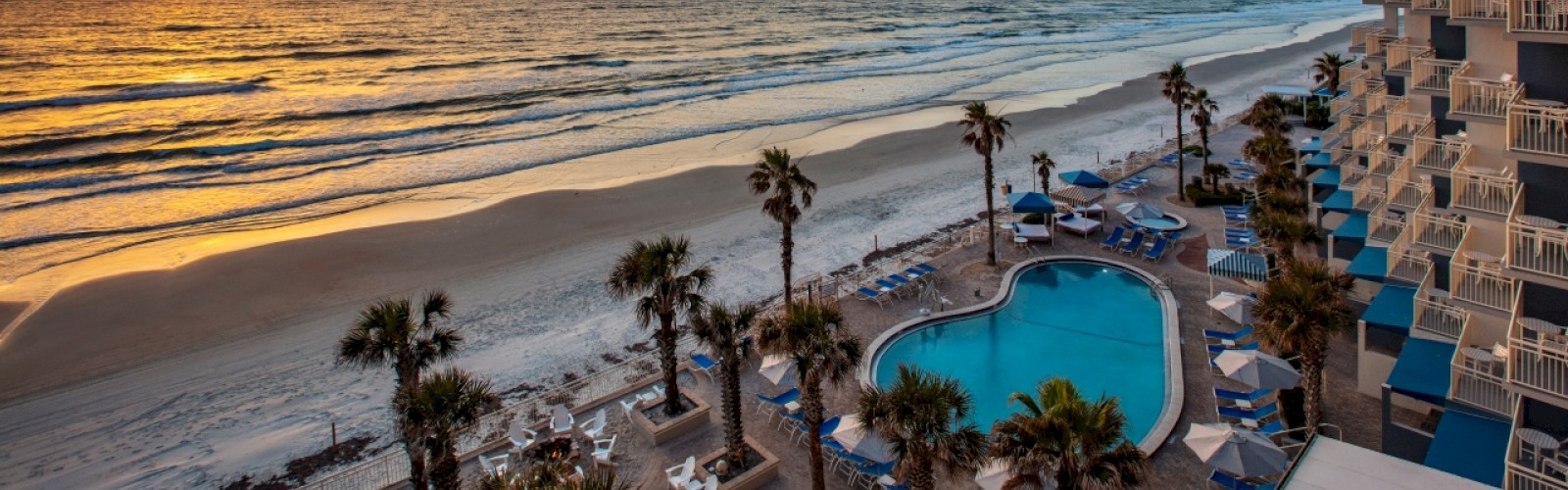 A beachside view with a pool, palm trees, and a vibrant sunset over the ocean, with a building nearby.