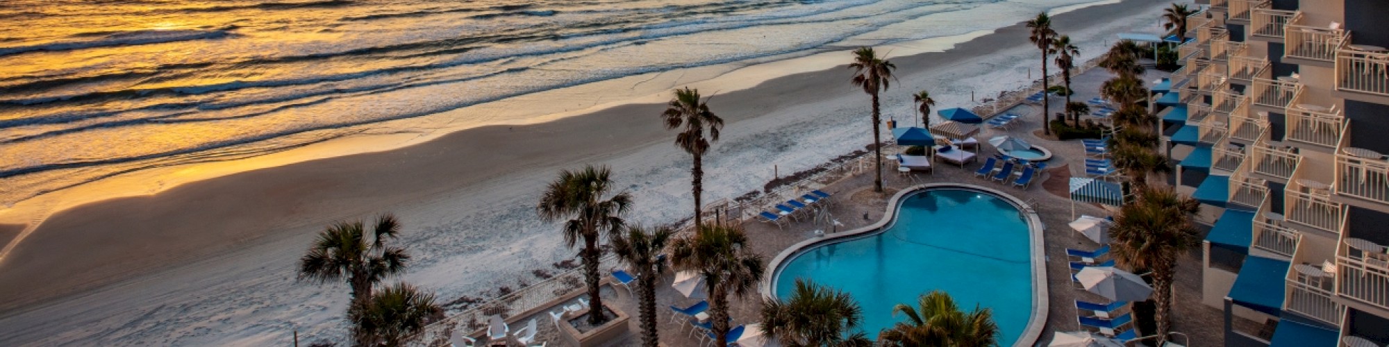 A beachside view with a pool, palm trees, and a vibrant sunset over the ocean, with a building nearby.