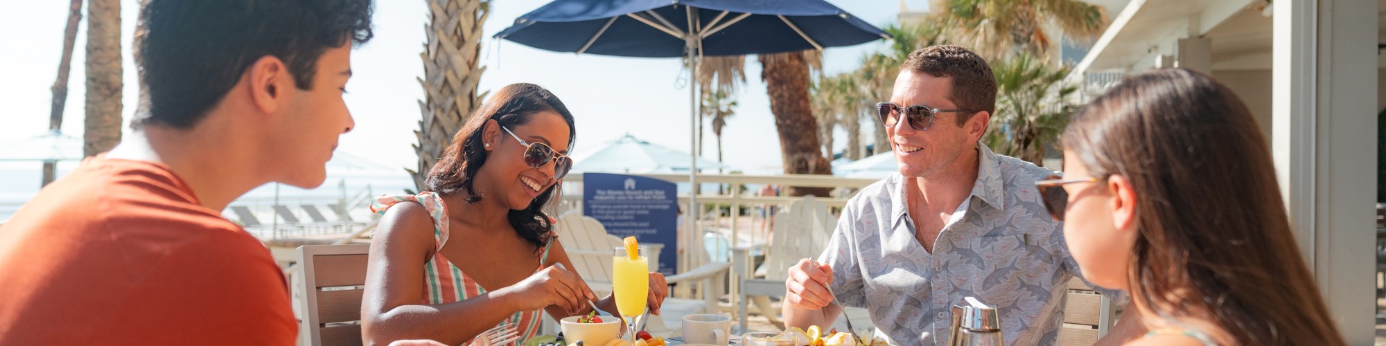 Four people are dining outdoors near palm trees, enjoying breakfast with juice and coffee at a beachfront location, creating a relaxed atmosphere.