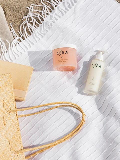 Beach-themed skincare products on a white towel with a straw bag, book, and a fringed cloth nearby at the beach.