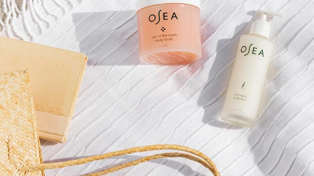 Beach-themed skincare products on a white towel with a straw bag, book, and a fringed cloth nearby at the beach.