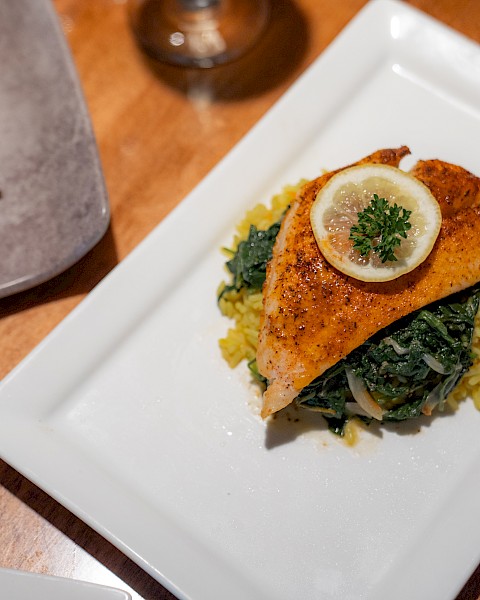 A plated dish featuring a piece of fish garnished with a lemon slice and parsley, served on a bed of spinach and yellow rice, alongside asparagus.