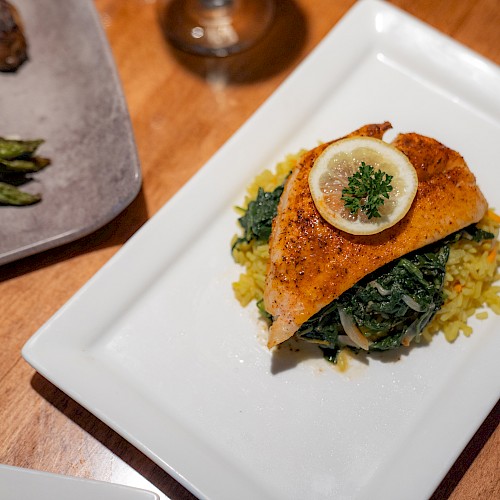 A plated dish featuring a piece of fish garnished with a lemon slice and parsley, served on a bed of spinach and yellow rice, alongside asparagus.