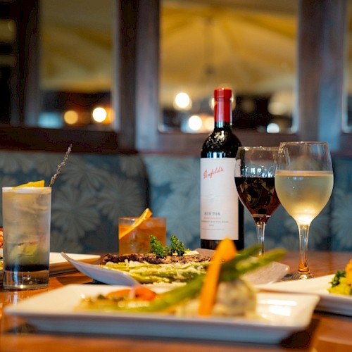 An elegant dinner setup featuring dishes, a bottle of wine, cocktails, and a glass of white wine on a wooden table.