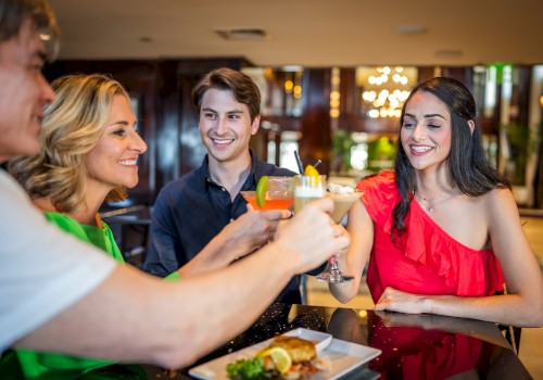 Four people are sitting around a table, enjoying drinks and food, smiling, and toasting each other.