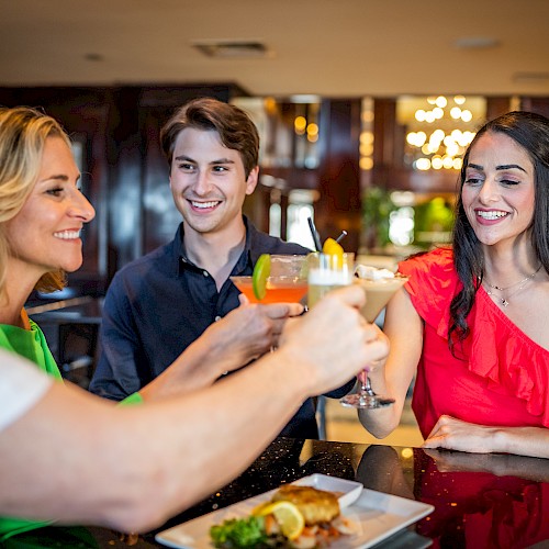 Four people are sitting around a table, enjoying drinks and food, smiling, and toasting each other.