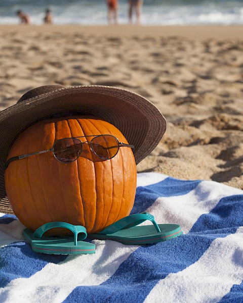 A pumpkin with sunglasses, a hat, and flip-flops sits on a beach towel with a toy bucket, at a sandy beach with people near the water.