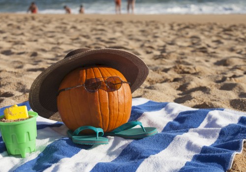 A pumpkin with sunglasses, a hat, and flip-flops sits on a beach towel with a toy bucket, at a sandy beach with people near the water.