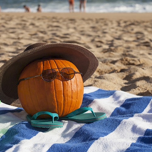 A pumpkin with sunglasses, a hat, and flip-flops sits on a beach towel with a toy bucket, at a sandy beach with people near the water.