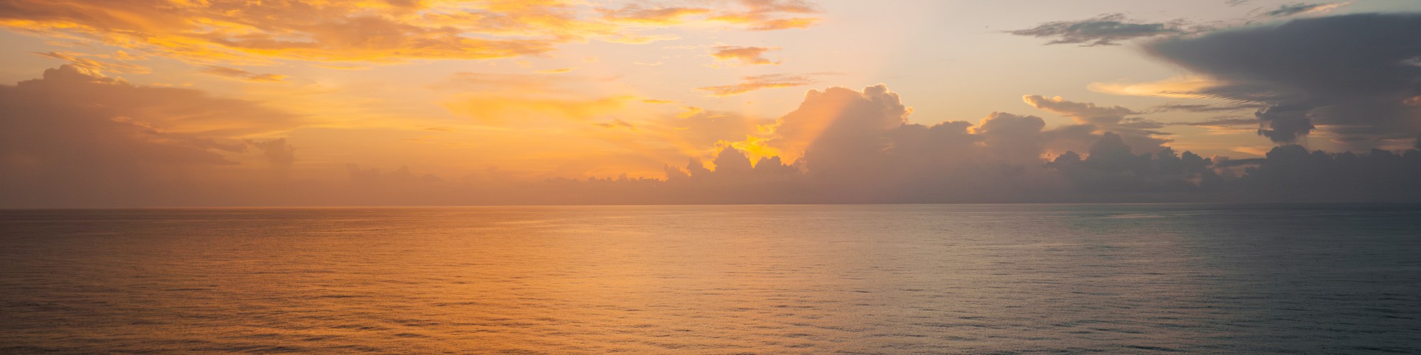 The image shows a serene ocean view at sunset, with a mix of orange, yellow, and dark clouds in the sky, reflecting on the calm waters.