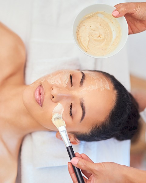 A person is receiving a facial treatment, with a face mask being applied by another person using a brush and a bowl of the mixture.