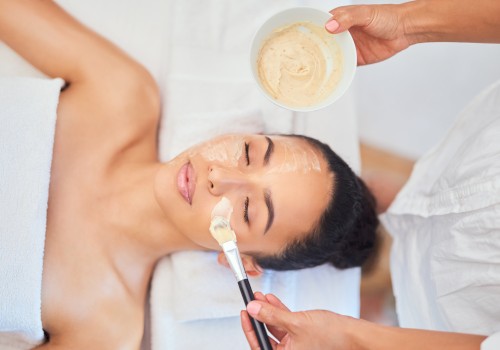 A person is receiving a facial treatment, with a face mask being applied by another person using a brush and a bowl of the mixture.