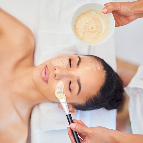 A person is receiving a facial treatment, with a face mask being applied by another person using a brush and a bowl of the mixture.