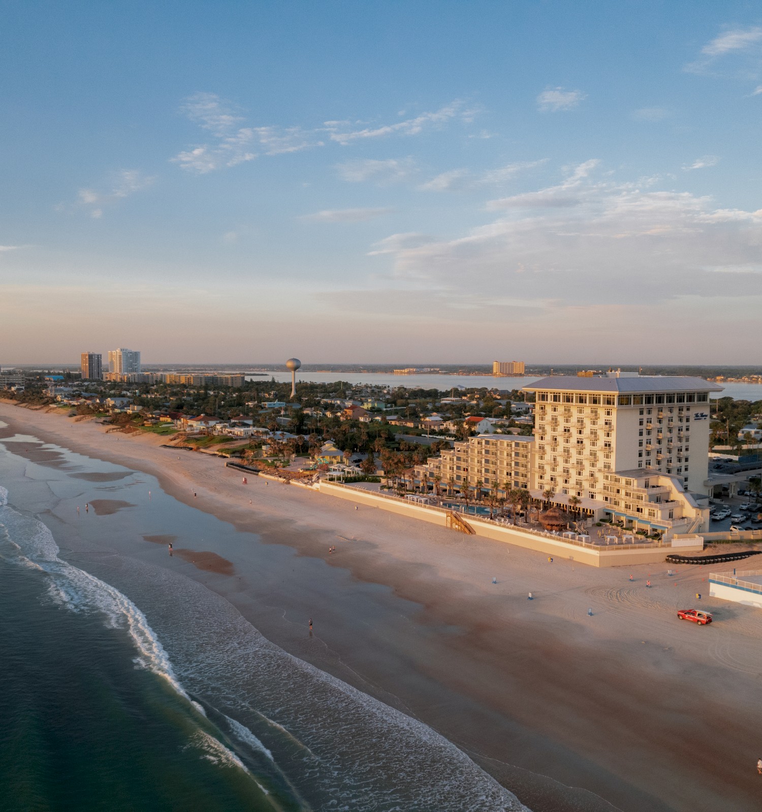 Aerial view of a coastal city with sandy beaches, buildings near the shoreline, and calm ocean waves under a partly cloudy sky during twilight.