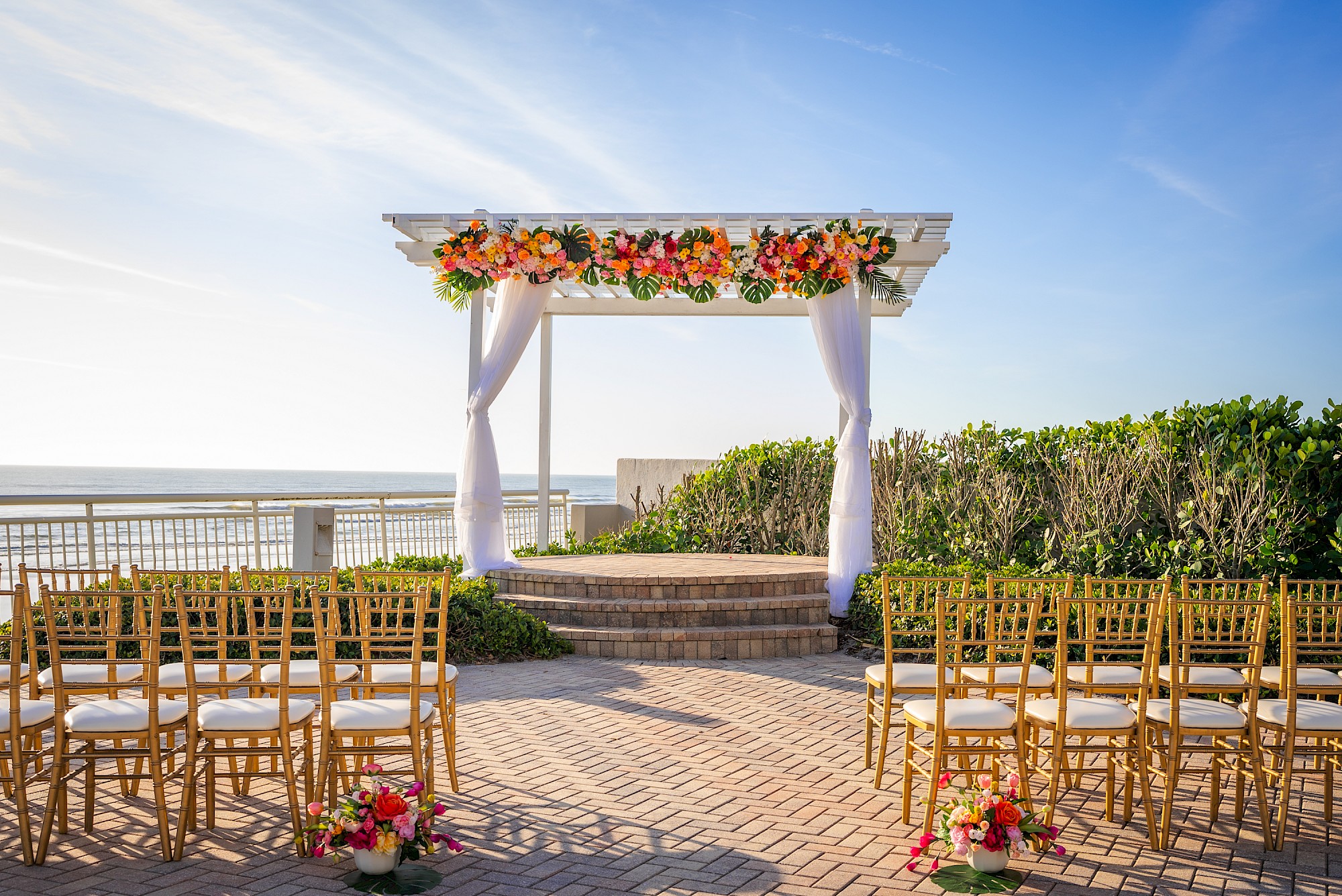 Outdoor wedding setup with gold chairs facing a decorated arch with flowers, under a clear blue sky near the ocean.