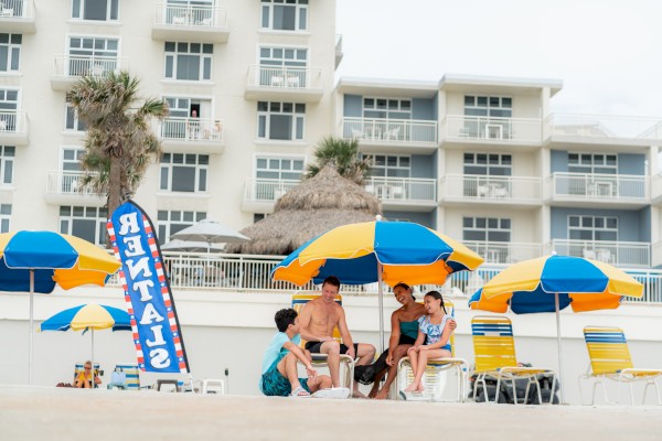 A group of people sit under colorful umbrellas on a sandy beach in front of a hotel building, with a sign reading 