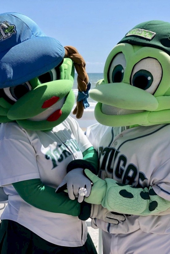 Two green mascots, one in a blue cap and the other in a green cap, pose together by a beach in playful outfits.