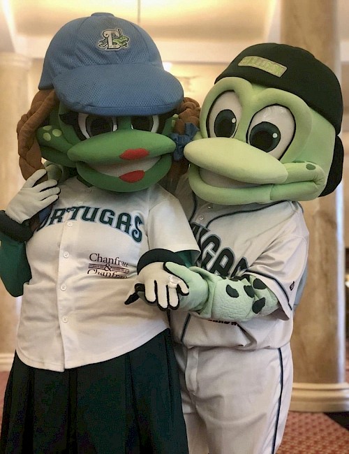 Two turtle mascots in baseball outfits pose together indoors, with one wearing a blue cap and the other a black cap.