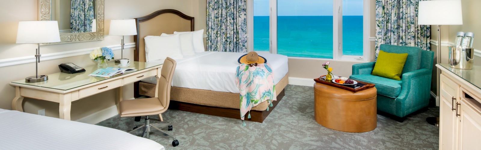 This image shows a hotel room with two beds, a desk, an armchair, and a window view of the ocean. The decor is modern yet cozy.